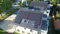 Das Bild zeigt zwei Gebäude mit großen Solarpanelen auf den Dächern. Die Panele sind gleichmäßig angeordnet und decken fast die gesamte Dachfläche ab. Um die Gebäude herum sind Bäume, eine Straße und einige Personen zu sehen, die sich auf einem Weg bewegen.