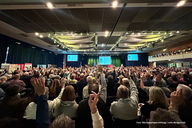 Eine große Menschenmenge in einem Konferenzsaal hebt die Hände. Im Hintergrund sind Bildschirme mit Informationen zu sehen. Die Stimmung ist aktiv und engagiert, was auf eine lebhafte Diskussion oder Abstimmung hindeutet.