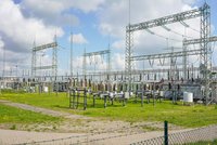 Das Bild zeigt eine Umspannstation mit mehreren elektrischen Anlagen und Transformatoren auf einer grünen Wiese. Hohe Metallgestelle mit Leitungen dominieren die Szene. Im Hintergrund sind einige Windkraftanlagen zu sehen, unter einem klaren Himmel mit wenigen Wolken.