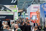 Die Aufnahme zeigt eine belebte Messehalle mit zahlreichen Besuchern, die zwischen verschiedenen Ständen flanieren. Im Hintergrund sind Werbeschilder und Stände von Unternehmen wie "reflex" und "STIEBEL" zu sehen. Die Atmosphäre wirkt geschäftig und informativ.