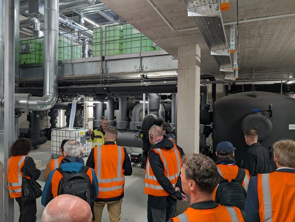Eine Gruppe von Personen in orangefarbenen Warnwesten hört einem Redner in einer Industrieumgebung zu. Im Hintergrund sind große schwarze Behälter und Rohre sichtbar. Die Atmosphäre wirkt informativ und industriell.