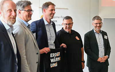 Fünf Männer stehen nebeneinander und lächeln. Sie halten ein schwarzes T-Shirt mit der Aufschrift "ERDERWÄRMUNG". Im Hintergrund befindet sich ein Banner mit dem Titel einer Veranstaltung. Die Umgebung wirkt professionell und förmlich.