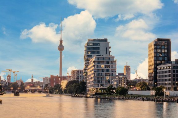 Blick auf die Berliner Skyline mit dem ikonischen Fernsehturm, der über modernen Gebäuden thront. Im Vordergrund fließt ein ruhiger Fluss und im Hintergrund sind Baukräne zu sehen, die auf städtische Entwicklungen hinweisen. Ein heller Himmel mit wenigen Wolken rundet das Bild ab.