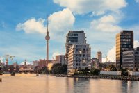 Blick auf die Berliner Skyline mit dem ikonischen Fernsehturm, der über modernen Gebäuden thront. Im Vordergrund fließt ein ruhiger Fluss und im Hintergrund sind Baukräne zu sehen, die auf städtische Entwicklungen hinweisen. Ein heller Himmel mit wenigen Wolken rundet das Bild ab.