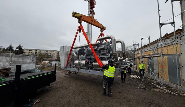 Ein Kran hebt einen großen Maschinenblock auf einer Baustelle. Arbeiter in gelben Sicherheitswesten sind in der Nähe aktiv, während die Maschine mit orangefarbenen Gurten gesichert wird. Im Hintergrund sind verschiedene Baustellenstrukturen sichtbar. Der Himmel ist grau und bewölkt.