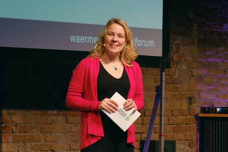 Eine lächelnde Frau steht auf einer Bühne, trägt eine pinkfarbene Jacke und ein schwarzes Kleid. Sie hält ein Blatt Papier in der Hand, während sie in die Kamera schaut. Im Hintergrund ist eine Steinmauer zu sehen.