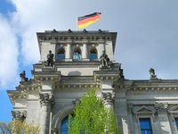Das Bild zeigt die Fassade eines historischen Gebäudes mit einer deutschen Flagge, die oben auf dem Dach weht. Auf den Giebeln sind mehrere Statuen sichtbar. Der Himmel im Hintergrund ist teilweise bewölkt, was dem Bild eine klare Atmosphäre verleiht.