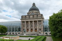 Das Bild zeigt ein historisches Gebäude mit einer großen Kuppel und Säulen an der Vorderseite. Vor dem Gebäude steht eine Statue eines Reiters. Im Hintergrund ist ein modernes Gewächshaus sichtbar, umgeben von gepflegten Gartenanlagen. Der Himmel ist bewölkt.