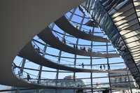 Das Bild zeigt die spiralförmige Innenansicht eines modernen Gebäudes mit transparenten Wänden. Viele Menschen gehen auf den spiralförmigen Treppen, die sich um den zentralen Raum schlingen, während Tageslicht durch die Fenster strömt und eine helle Atmosphäre schafft.