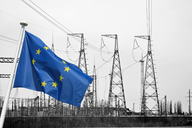 Die europäische Flagge weht im Vordergrund vor einer schwarz-weißen Landschaft mit Hochspannungsmasten im Hintergrund. Die Masten sind in verschiedenen Höhen und Formen dargestellt, was die industrielle Umgebung betont.