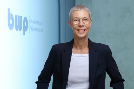 Eine lächelnde Frau mit kurzem, grauen Haar trägt eine Brille und einen blauen Blazer. Sie steht vor einer hellen Wand mit dem Logo und Namen des Bundesverbands Wärmepumpe e.V. Die Atmosphäre ist freundlich und professionell.