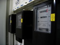 Das Bild zeigt mehrere schwarze Stromzähler, die nebeneinander in einem Schaltkasten montiert sind. Auf den Zählern sind digitale Anzeigen sichtbar, die den Energieverbrauch in Kilowattstunden anzeigen. Die Zähler sind mit gelben Aufklebern gekennzeichnet.