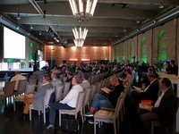 Ein großer Saal mit einer Vielzahl von Personen, die in Stuhlreihen sitzen und einem Vortrag folgen. Auf der Bühne steht ein Redner, während ein Bildschirm hinter ihm Informationen zeigt. Das Ambiente ist modern mit Steinen und grünen Lichtern an den Wänden.