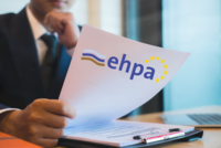 Eine Person in formeller Kleidung hält ein Dokument in der Hand, auf dem das Logo der EHPA und die EU-Flagge abgebildet sind. Im Hintergrund sind verschwommene Fenster und ein Tisch mit einem Stift zu sehen.