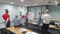 Eine Gruppe von fünf Personen steht in einem Besprechungsraum um einen Flipchart. Eine Person erklärt etwas, während die anderen aufmerksam zuhören. Auf einem Tisch sind Laptops und Unterlagen verteilt. Der Raum hat große Fenster und einfache Möbel.