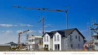 Die Abbildung zeigt eine Baustelle mit mehreren Kränen und einem gerade fertiggestellten weißen Wohnhaus. Im Hintergrund sind Gerüste und weitere Bauarbeiten zu sehen, unter einem klaren, blauen Himmel. Es vermittelt den Eindruck einer aktiven Bauumgebung.
