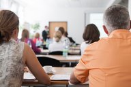 Eine Gruppe von Menschen sitzt an Tischen in einem Seminarraum. Im Vordergrund sind zwei Personen von hinten zu sehen. Im Hintergrund spricht ein Dozent, während auf einer Leinwand Informationen präsentiert werden. Wasserflaschen und Tassen stehen auf den Tischen.