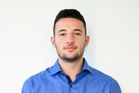 Ein junger Mann mit kurzen, dunklen Haaren und einem gepflegten Bart schaut direkt in die Kamera. Er trägt ein blaues Hemd und steht vor einem hellen Hintergrund. Sein Ausdruck wirkt freundlich und selbstbewusst.