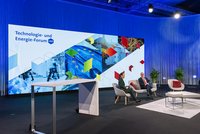 Das Bild zeigt ein modernes Podium mit zwei Sprechern, die auf einem beigen Teppich in eleganten Sesseln sitzen. Im Hintergrund ist eine große Grafik mit dem Titel „Technologie- und Energie-Forum LIVE“ sowie dynamischen, grafischen Elementen und einer EU-Flagge zu sehen.