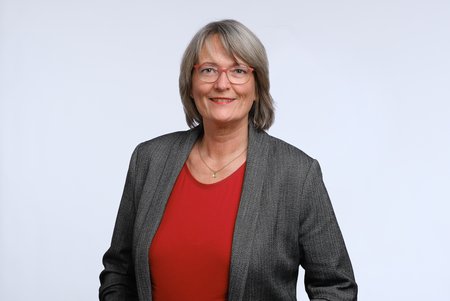 Eine lächelnde Frau steht vor einem einfarbigen Hintergrund. Sie trägt eine graue Jacke über einem roten Oberteil. Ihre Haare sind schulterlang und grau, und sie hat eine Brille mit einem dünnen Rahmen. Die Frau wirkt freundlich und selbstbewusst.