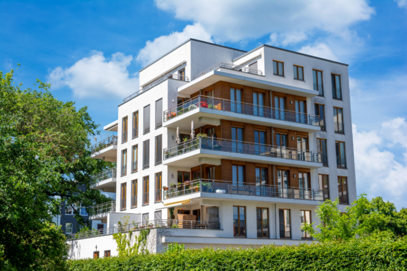 Ein modernes Wohngebäude mit mehreren Etagen und Balkonen, umgeben von Bäumen und Sträuchern. Die Fassade ist hell und weist große Fenster auf, die viel Licht hereinlassen. Der Himmel ist blau mit einigen Wolken.