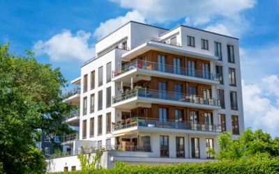 Ein modernes Wohngebäude mit mehreren Etagen und Balkonen, umgeben von Bäumen und Sträuchern. Die Fassade ist hell und weist große Fenster auf, die viel Licht hereinlassen. Der Himmel ist blau mit einigen Wolken.