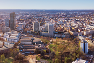 Luftaufnahme einer Stadt, die moderne Hochhäuser und historische Gebäude zeigt. Im Vordergrund sind grüne Parkanlagen sichtbar. Die Skyline erstreckt sich über eine weite Fläche, mit einer klaren Sicht auf die städtische Umgebung und den blauen Himmel.