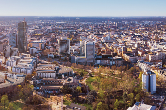 Luftaufnahme einer Stadt, die moderne Hochhäuser und historische Gebäude zeigt. Im Vordergrund sind grüne Parkanlagen sichtbar. Die Skyline erstreckt sich über eine weite Fläche, mit einer klaren Sicht auf die städtische Umgebung und den blauen Himmel.