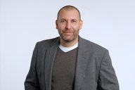 Ein Mann mit kurzen Haaren und Bart trägt einen grauen Blazer über einem grauen Pullover und einem weißen Shirt. Er steht vor einem einfarbigen, hellen Hintergrund und schaut direkt in die Kamera.