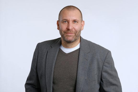 Ein Mann mit kurzen Haaren und Bart trägt einen grauen Blazer über einem grauen Pullover und einem weißen Shirt. Er steht vor einem einfarbigen, hellen Hintergrund und schaut direkt in die Kamera.