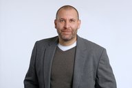 Ein Mann mit kurzen Haaren und Bart trägt einen grauen Blazer über einem grauen Pullover und einem weißen Shirt. Er steht vor einem einfarbigen, hellen Hintergrund und schaut direkt in die Kamera.