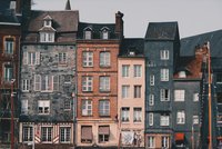 Eine Reihe von historischen Gebäuden mit unterschiedlichen Farben und Materialien, darunter Ziegel und Schiefer, an einer Hafenmauer. Einige Fenster sind geöffnet, andere haben Jalousien. Im Vordergrund sind Bootsmasten zu sehen, die zur maritimen Atmosphäre beitragen.