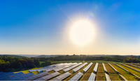 Eine große Solaranlage mit zahlreichen Solarzellen erstreckt sich über ein Feld, beleuchtet von einer strahlenden Sonne. Der Himmel ist klar und blau, während im Hintergrund grüne Bäume und sanfte Hügel sichtbar sind. Die Szene vermittelt ein Gefühl von Nachhaltigkeit und erneuerbarer Energie.