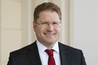 Porträt eines Mannes mit Brille, der in einem dunklen Anzug und roter Krawatte lächelt. Der Hintergrund ist hell und neutral, was den Fokus auf sein freundliches Gesicht lenkt.