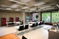 Ein moderner Besprechungsraum mit weißen Tischen und schwarzen Stühlen. An der Wand hängen drei farbige Bilder mit floralem Muster. In der Ecke steht ein großer Bildschirm, und durch das Fenster ist ein grüner Außenbereich sichtbar.