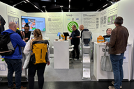 Ein Messestand mit mehreren Personen, die sich um einen Tisch versammeln. Auf dem Tisch befinden sich Geräte und Informationsmaterialien. Im Hintergrund sind Werbematerialien und Grafiken zur Firma "bwp" zu sehen. Die Atmosphäre ist lebhaft und informativ.