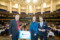 Drei Personen stehen auf einer Bühne in einem großen Veranstaltungsraum. Der Hintergrund zeigt eine große Menge von Zuhörern an Tischen. Ein Schild mit der Aufschrift "Wärmepumpen Initiative" ist sichtbar. Die Atmosphäre vermittelt eine formelle Veranstaltung oder Konferenz.