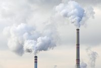 Zwei hohe Schornsteine sind im Bild zu sehen, aus denen dichter, weißer Rauch aufsteigt. Der Himmel wirkt grau und bewölkt, was die verschmutzte Atmosphäre unterstreicht. Die Schornsteine symbolisieren industrielle Emissionen und Umweltbelastung.