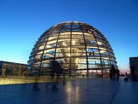Eine gläserne Kuppel der Reichstagsgebäude in Berlin bei Dämmerung. Die Struktur ist in einem modernen Stil gestaltet und leuchtet von innen. Einige Personen gehen auf dem Platz vor dem Gebäude, während der Himmel in sanften Blau- und Lilatönen schimmert.
