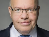 Porträt eines älteren Mannes mit Brille, der direkt in die Kamera schaut. Er trägt einen dunklen Anzug und ein helles Hemd. Sein Gesicht zeigt ein freundliches Lächeln, während der Hintergrund einheitlich grau ist.