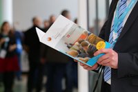 Ein Mann in einem Anzug hält ein Faltblatt des Kongresses 2016, das Informationen über Veranstaltungen und Programme enthält. Im Hintergrund sind verschwommene Personen zu sehen, die an der Veranstaltung teilnehmen.