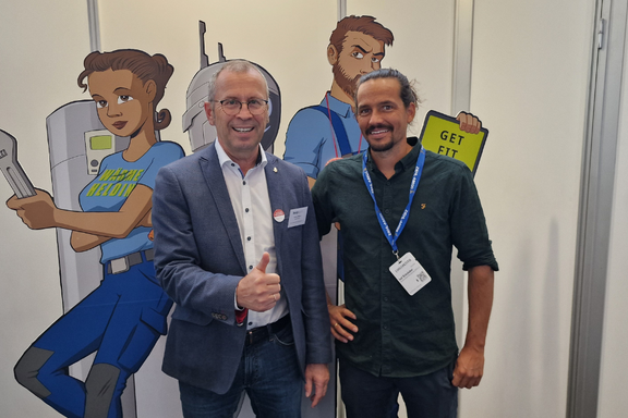 Zwei Erwachsene stehen vorne vor einer Wand mit Comicfiguren. Der Mann links trägt einen Anzug und zeigt Daumen hoch, während der Mann rechts in einem dunklen Hemd lächelt. Im Hintergrund sind farbige Zeichnungen von Charakteren zu sehen, die in einem technischen Umfeld arbeiten.