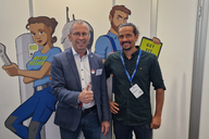 Zwei Erwachsene stehen vorne vor einer Wand mit Comicfiguren. Der Mann links trägt einen Anzug und zeigt Daumen hoch, während der Mann rechts in einem dunklen Hemd lächelt. Im Hintergrund sind farbige Zeichnungen von Charakteren zu sehen, die in einem technischen Umfeld arbeiten.