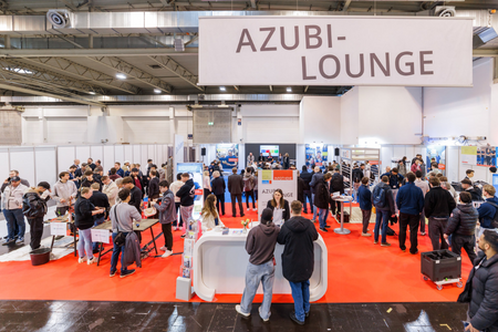 Eine lebhafte Messe mit vielen Menschen steht im Mittelpunkt. In der Mitte befindet sich ein Empfangstisch mit dem Schild "AZUBI-LOUNGE". Besucher interagieren und informieren sich, während Aussteller in einem großen, hellen Raum ihre Angebote präsentieren. Der Boden ist mit rotem Teppich ausgelegt.