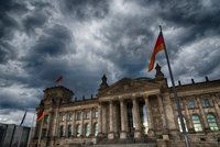 Das Bild zeigt das Reichstagsgebäude in Berlin unter einem dramatischen, bewölkten Himmel. Die Fassade ist ???????, mit Säulen und großen Fenstern. Mehrere Fahnen, darunter die deutsche Flagge, wehen an den Masten. Die Atmosphäre wirkt eindrucksvoll und imposant.