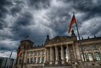 Das Bild zeigt das Reichstagsgebäude in Berlin unter einem dramatischen, bewölkten Himmel. Die Fassade ist ???????, mit Säulen und großen Fenstern. Mehrere Fahnen, darunter die deutsche Flagge, wehen an den Masten. Die Atmosphäre wirkt eindrucksvoll und imposant.