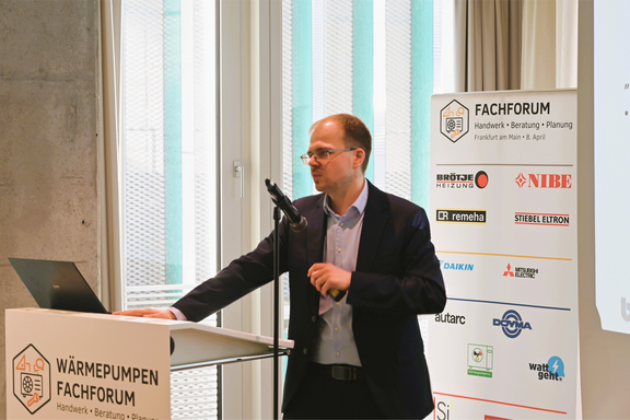Ein Referent steht an einem Rednerpult während des Wärmepumpen Fachforums. Er spricht, während Informationen im Hintergrund und Logos von Sponsoren zu sehen sind. Großen Fenstern im Hintergrund lassen viel Licht hinein.