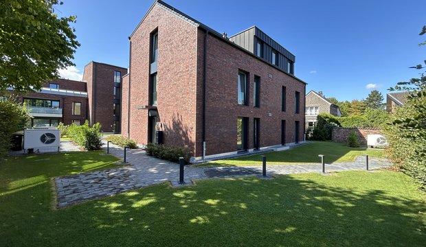 Ein modernes, rotbraunes Wohngebäude mit großen Fenstern und einem Flachdach. Im Vordergrund führt ein gepflasterter Weg durch eine gepflegte Grünfläche. Die Szenerie ist bei klarem, sonnigem Wetter aufgenommen, und Bäume sind im Hintergrund sichtbar.