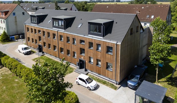 Ein modernes, mehrstöckiges Wohngebäude mit Holzfassade ist im Vordergrund zu sehen. Es verfügt über mehrere Fenster und Eingänge. Vor dem Gebäude stehen verschiedene Autos. Im Hintergrund sind weitere Wohngebäude und Bäume zu erkennen. Der Himmel ist klar und blau.