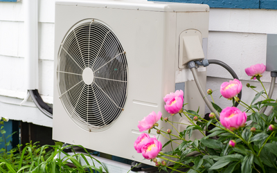 Klimaanlage an einer Außenwand, umgeben von rosa Pfingstrosen. Die Einheit hat einen großen Ventilator und ist in einem freundlichen, gepflegten Garten platziert.